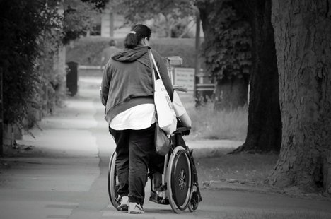 Die schwarz-weiß-Fotografie zeigt eine Frau von hinten, die einen Rollstuhl schiebt.
